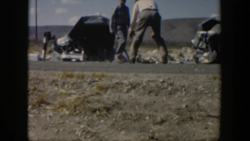 1951 Texas 8mm frame showing men near an overturned dark sedan and scattered wreckage on a rural roadside, with low hills or open land behind them.