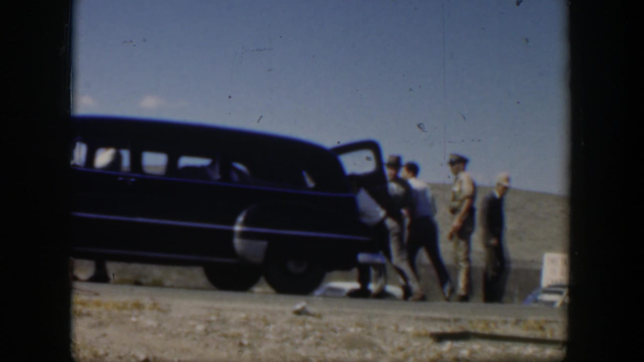 1951 Texas home-movie frame showing men gathered by a dark mid-century station wagon on a gravel roadside under a blue sky.