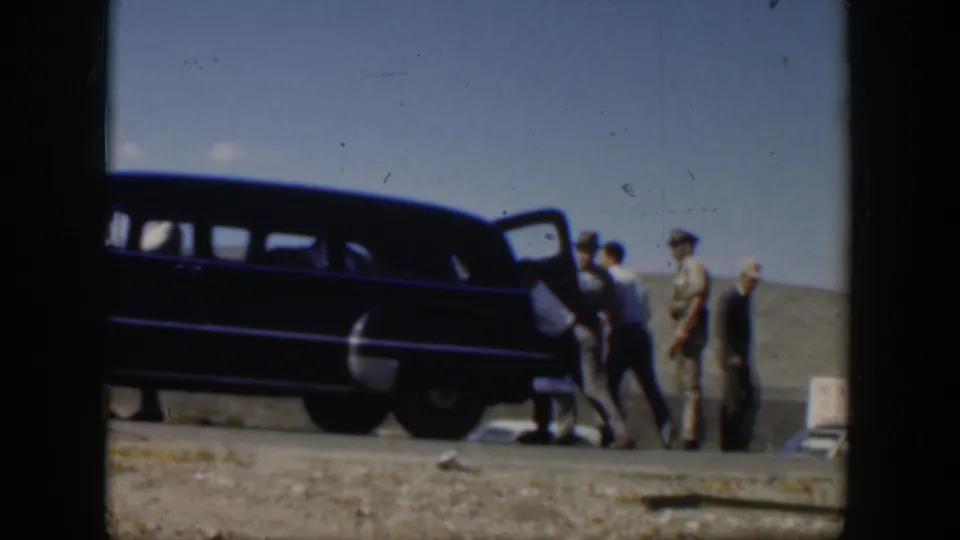 1951 Texas home-movie frame showing men gathered by a dark mid-century station wagon on a gravel roadside under a blue sky.