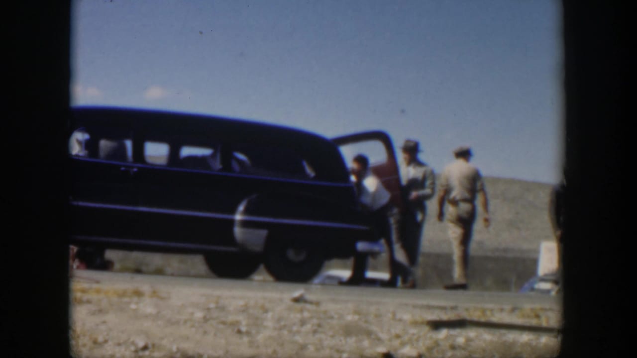 1951 Texas roadside frame with a man bending near an open vehicle door as other men watch beside a dark station wagon.