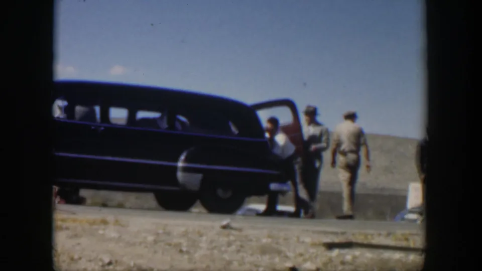 1951 Texas roadside frame with a man bending near an open vehicle door as other men watch beside a dark station wagon.