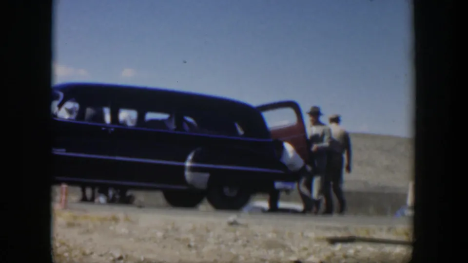 1951 Texas 8mm frame showing several men close to the open side of a large dark station wagon on a dusty roadside.