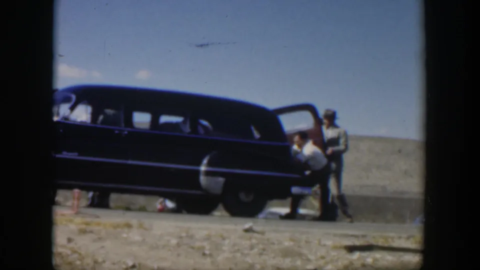 1951 Texas frame showing a man bent beside a dark station wagon as another man stands at the open door on a dry roadside.