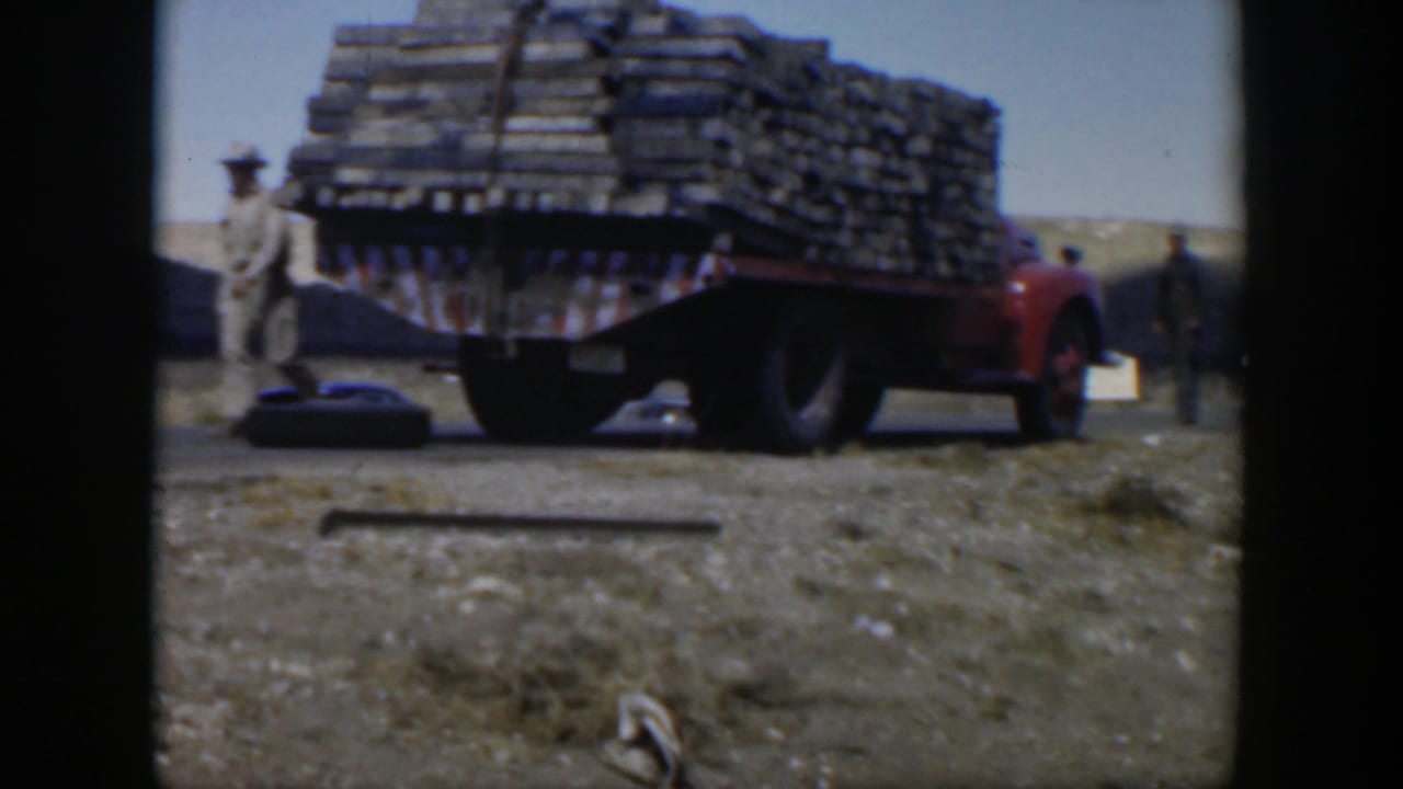 Red truck loaded with wooden planks beside dusty roadside with a man in light clothing nearby.