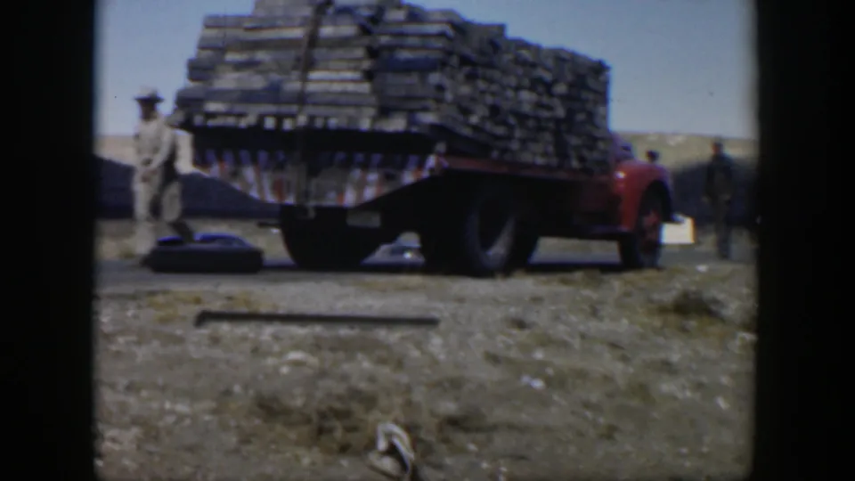 1951 Texas home-movie frame showing a red truck stacked with wooden planks on dusty ground, with a man walking at left.