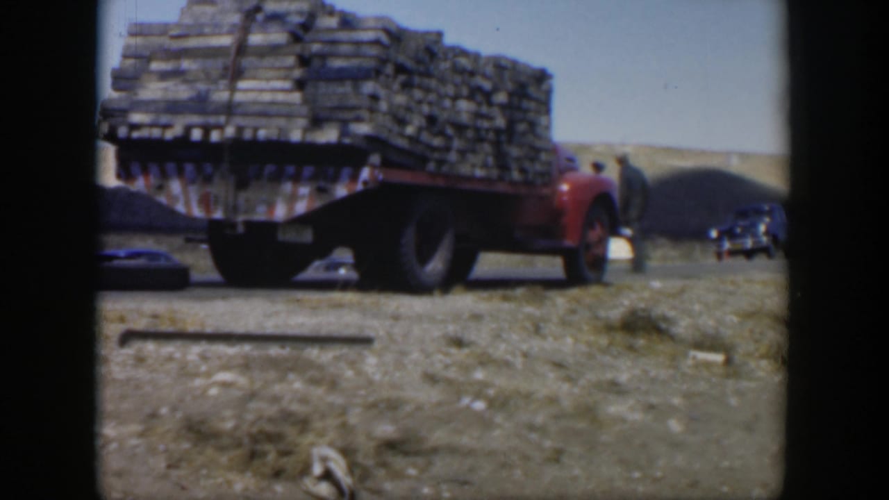 The red plank-loaded truck remains foregrounded in a Texas roadside scene, with adult figures and dark cars beyond it.