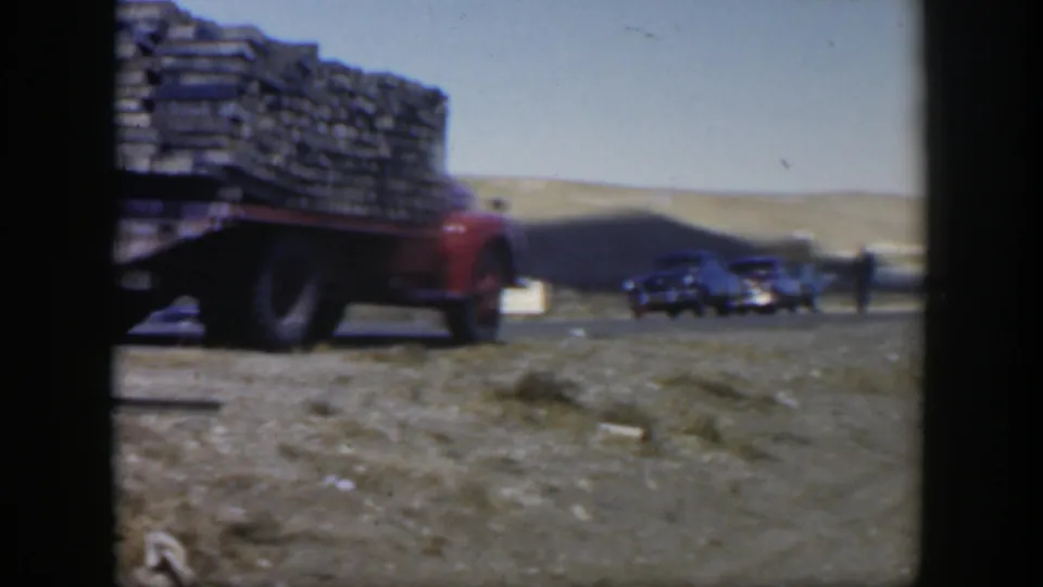 A soft 8mm frame shows the plank-loaded red truck at left and several dark cars on the roadside in 1951 Texas.
