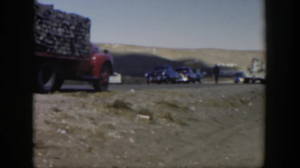 Wide 8mm view of the red truck at left, dark cars, and small adult figures on a clear Texas roadside in 1951.