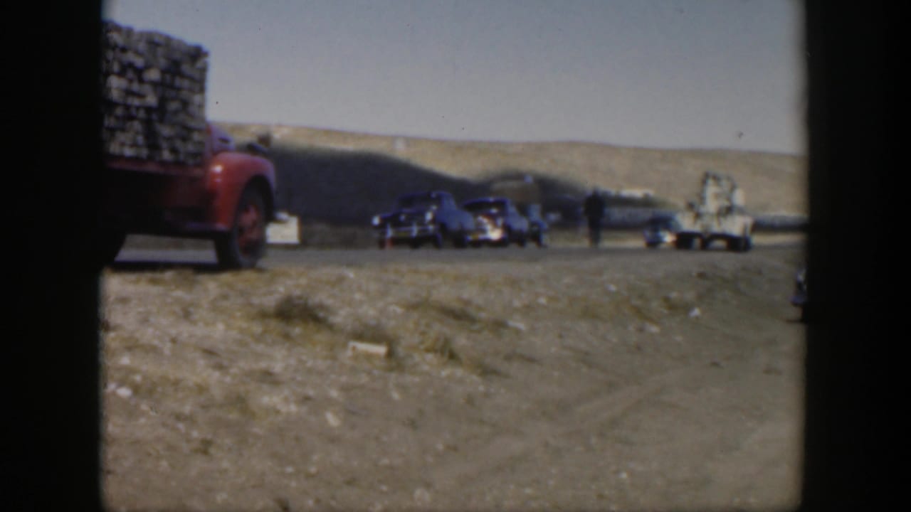 Red loaded truck at left with classic cars and people stopped on a dusty rural road.