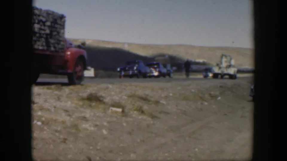 1951 Texas home-movie frame with a red loaded truck, several classic cars, and people on a dusty rural road below low hills.