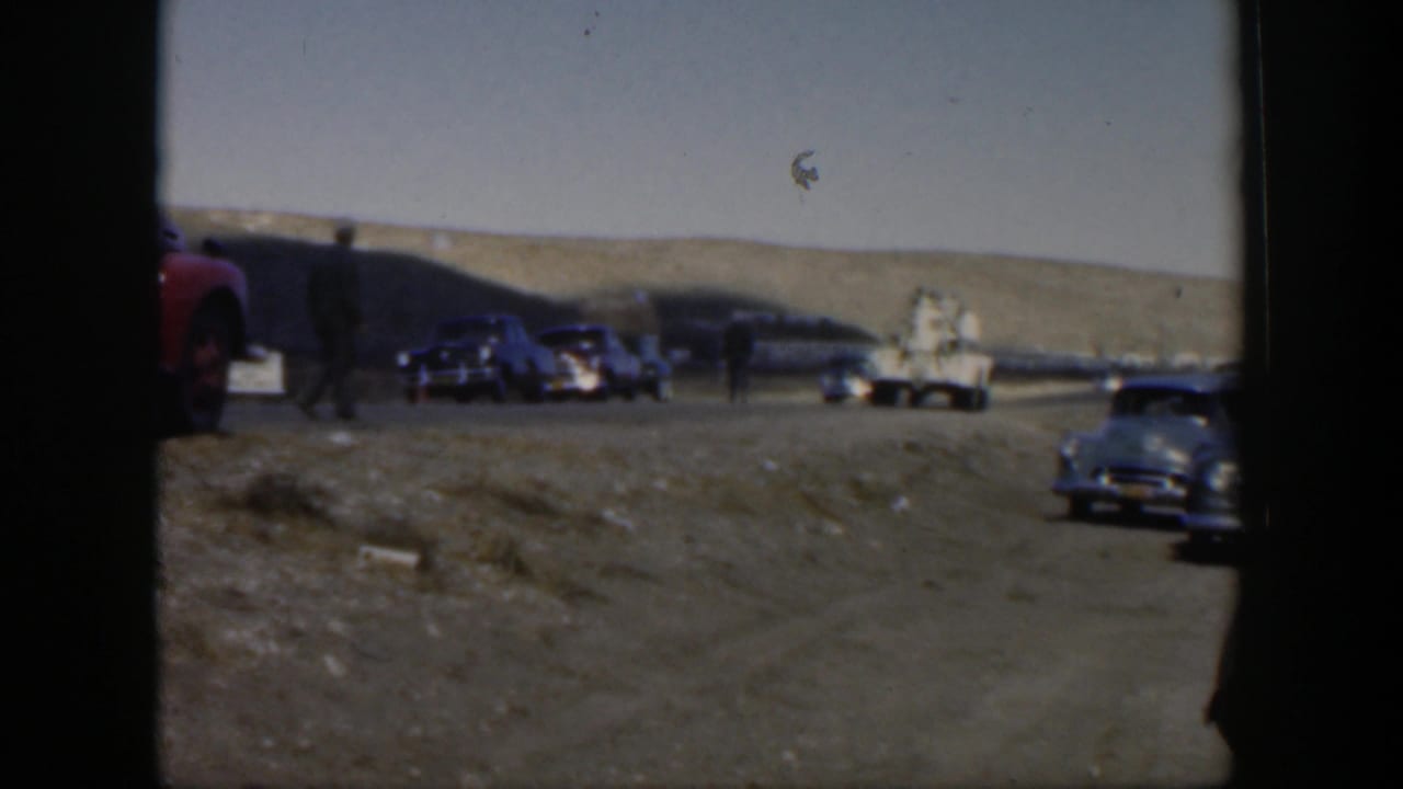 1951 Texas roadside view with dark sedans, a light utility-style truck, and a person walking across the dusty open road.