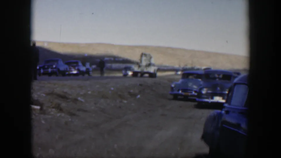 1951 Texas home-movie frame showing stopped classic cars, a pale truck in the distance, and a dark sedan close to the camera.