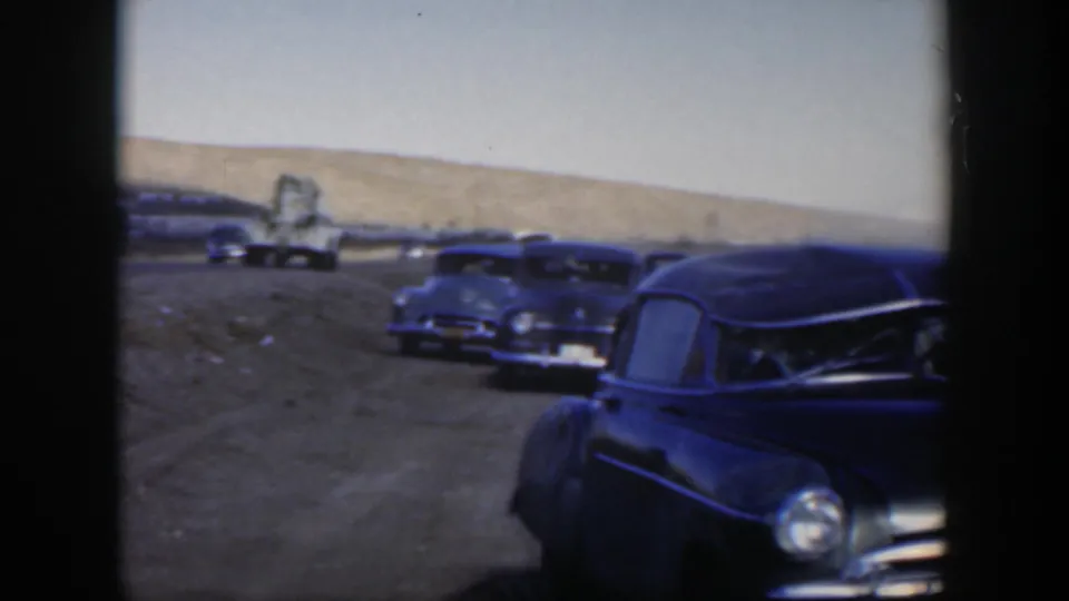 1951 Texas rural road frame with dark vintage cars in the foreground and several stopped vehicles farther along the road.