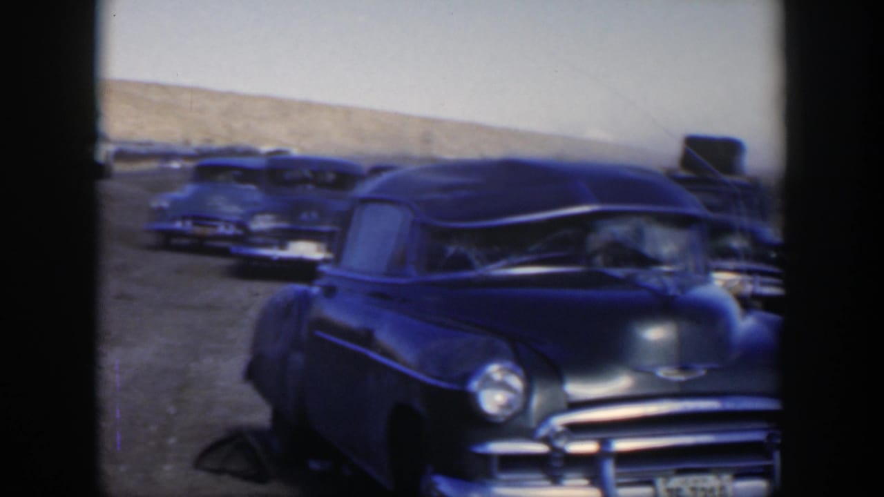 1951 Texas 8mm frame showing a blue sedan with chrome trim parked among other cars on open dusty ground.