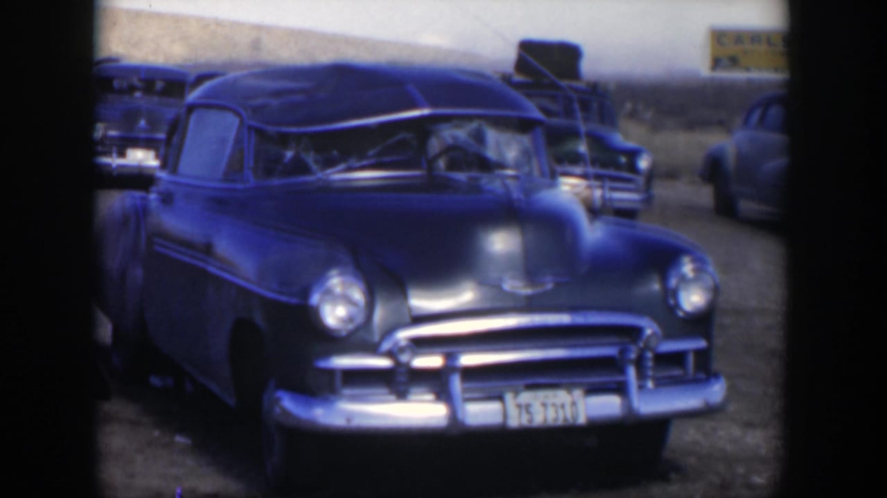1951 Texas frame with a blue sedan facing the camera, other vehicles nearby, and a partially legible roadside sign behind it.