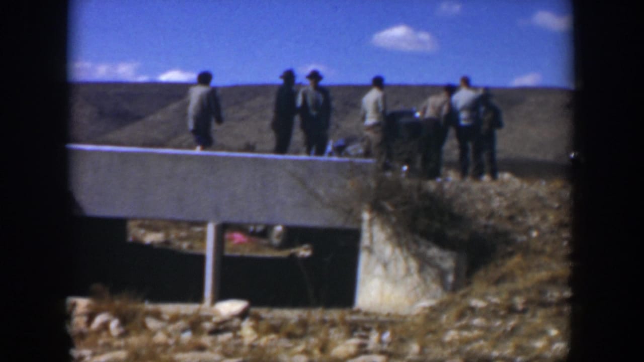 Men stand near a damaged green car beside a concrete roadside structure.