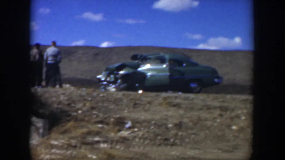 1951 Texas 8mm frame showing a damaged green car parked on tan ground with two men partly visible nearby.