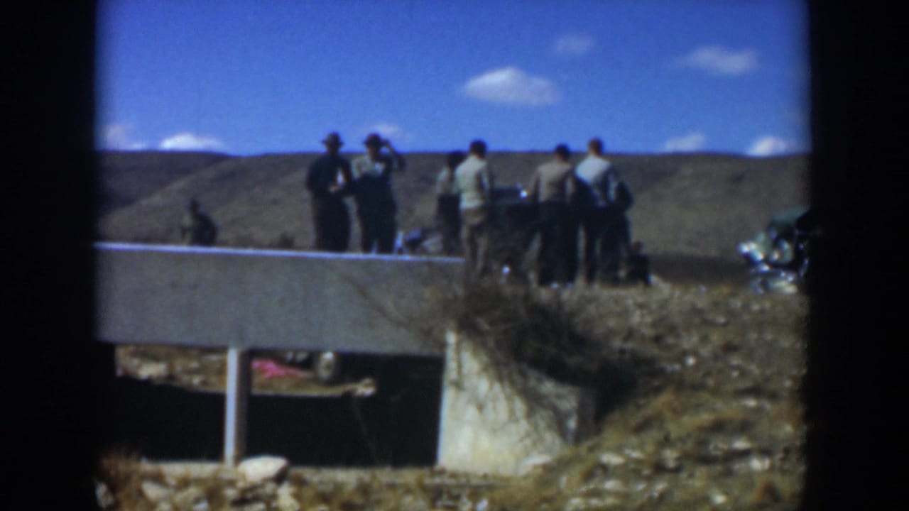 Small group of people standing outdoors near a low concrete wall under a blue sky.