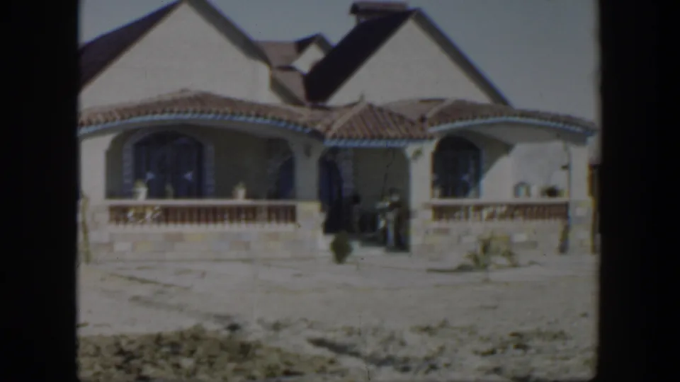 1951 Texas home-movie frame showing the front of a stucco house with arched windows, tiled roof, porch railing, and sparse yard.