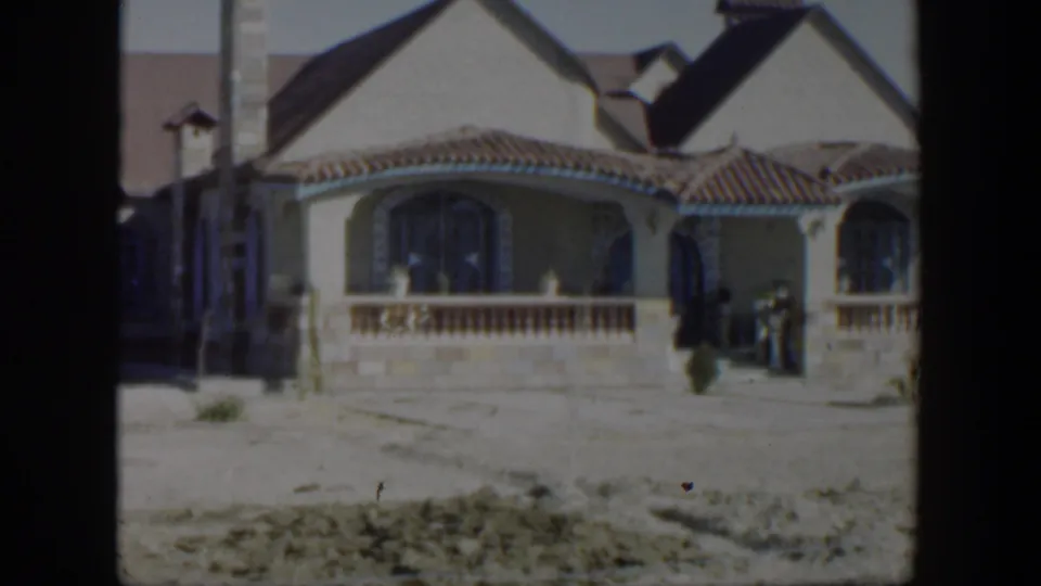 1951 Texas frame of a stucco residential exterior with arched window, tiled roof, porch railing, and dry foreground soil.