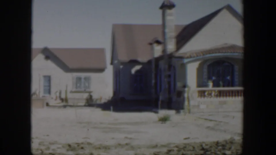 1951 Texas 8mm frame showing a smaller neighboring house beside the larger stucco home, with bright sun and a sparse yard.