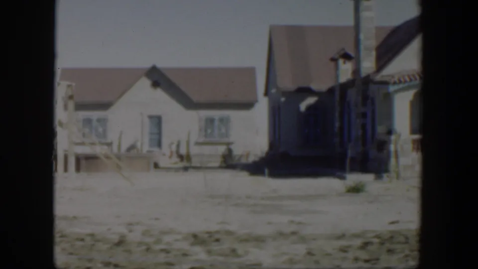 1951 Texas home-movie frame showing the smaller house and part of the larger stucco house across a barren, sunlit yard.