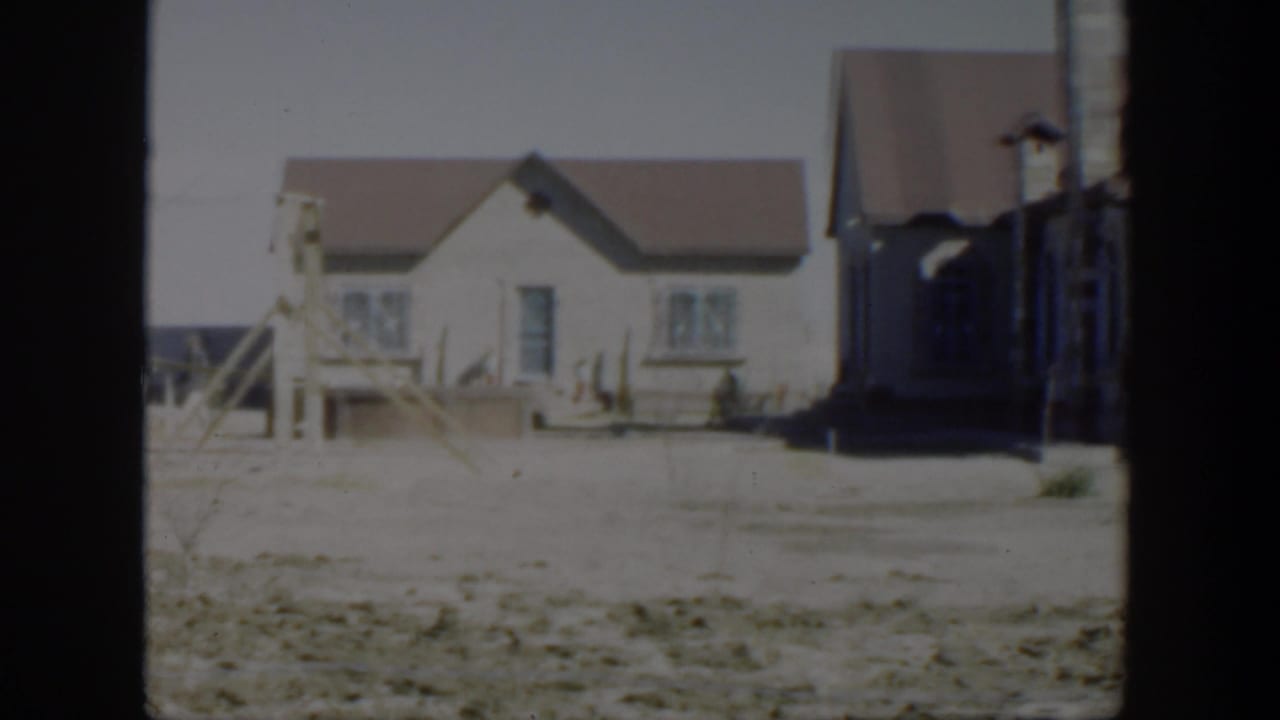 White-sided house with a brown roof beyond a dry sandy foreground.