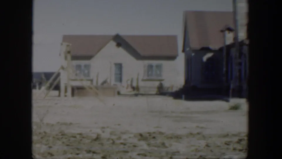 1951 Texas home-movie frame showing a white-sided house, brown pitched roof, bare yard, and sparse fencing in a dry residential setting.