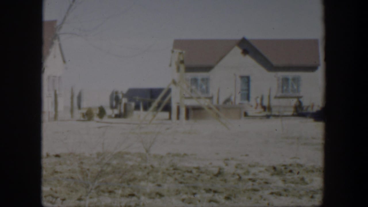 1951 Texas residential exterior with a small white house, porch steps, fence posts, dry ground, and a dark parked vehicle nearby.