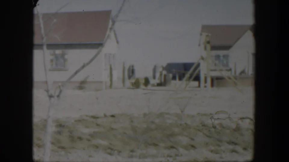 1951 Texas home-movie view of a residential house partly obscured by bare branches, with fence posts and dry open ground in front.