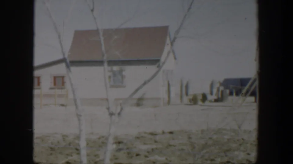 1951 Texas residential frame showing the side of a white house, brown roof, bare tree branches, fence posts, and a dark vehicle in the background.