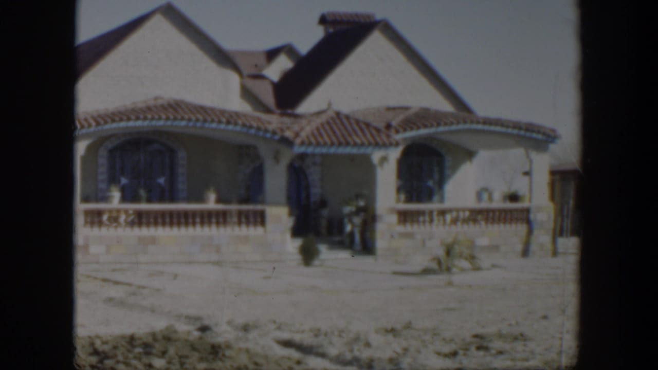 House front with arched windows, tiled roof, porch, dry yard, and small figure near entrance.