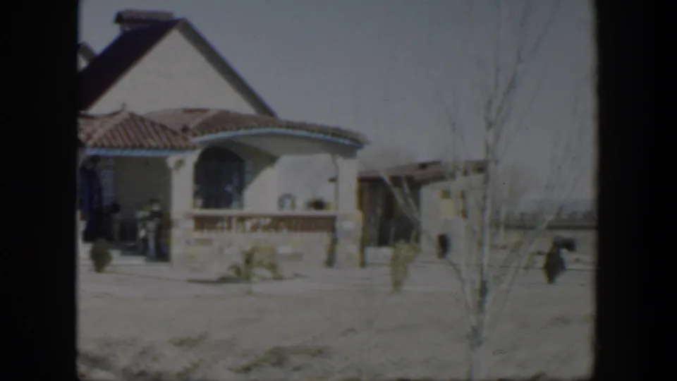 Late frame of the 1951 Texas house exterior, showing the arched porch, tiled roof, sparse yard, bare tree, and adjacent structure.