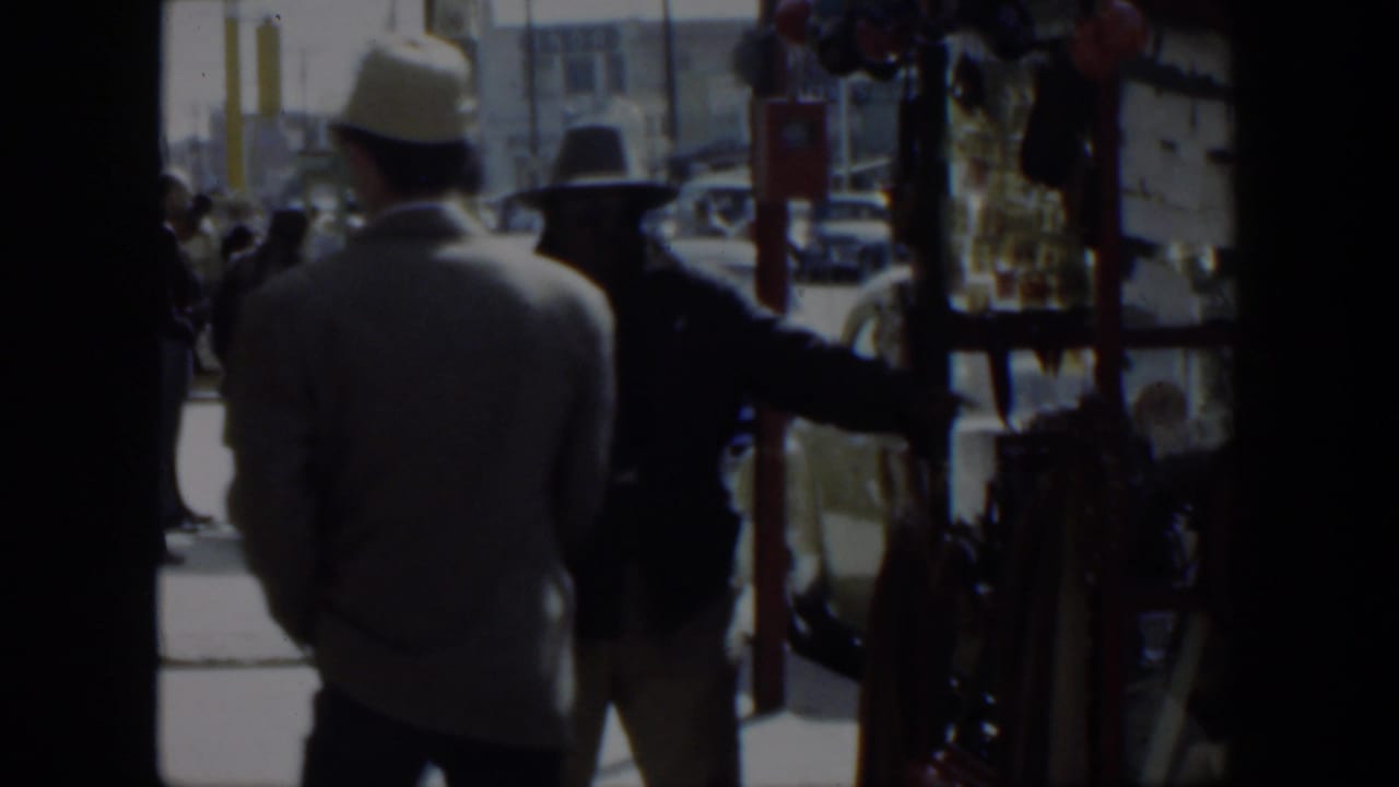 1951 Texas 8mm frame showing two men in hats near a shop entrance, with cars and a red stall visible outside.