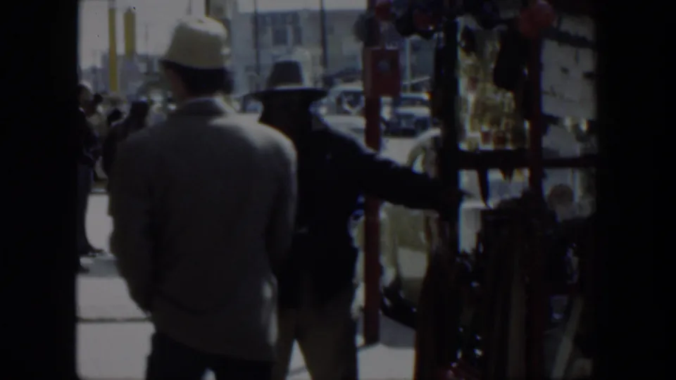 1951 Texas 8mm frame showing two men in hats near a shop entrance, with cars and a red stall visible outside.