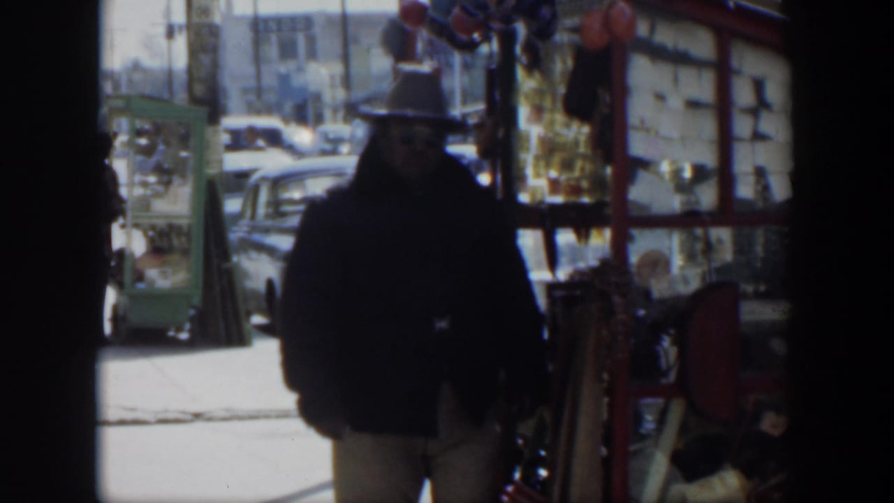 1951 Texas street frame with a man in a dark jacket and hat walking past a red stall with balloons and cars behind him.