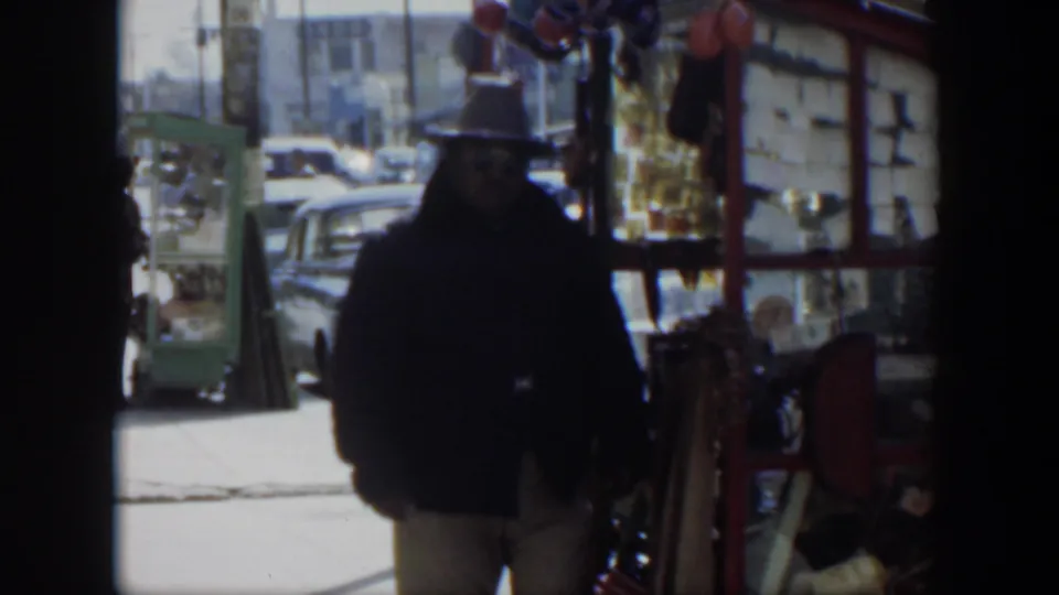 1951 Texas street frame with a man in a dark jacket and hat walking past a red stall with balloons and cars behind him.