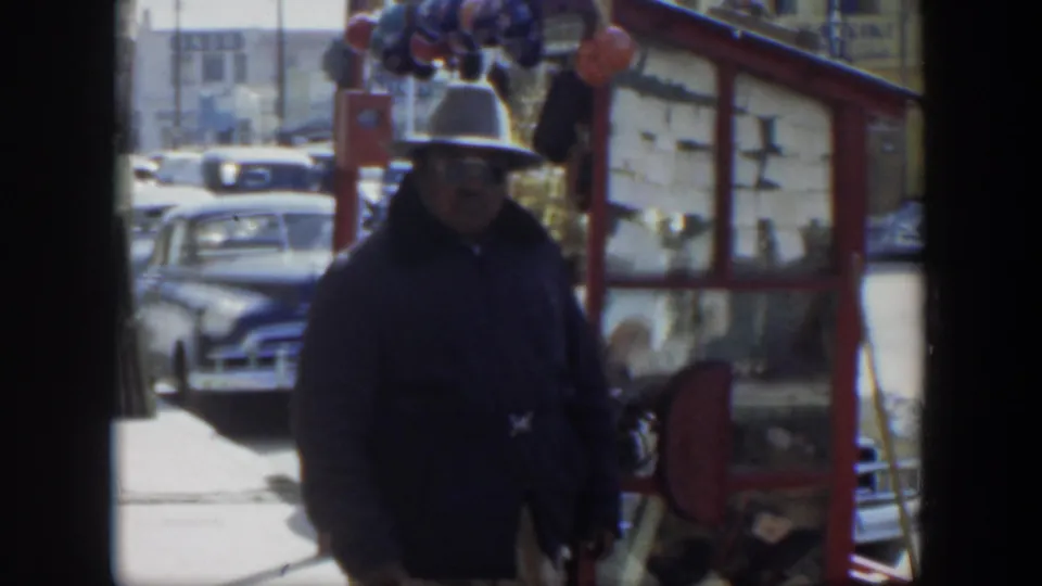 1951 Texas 8mm frame of a man in a hat and dark jacket walking near a red street stall with mid-century cars behind.