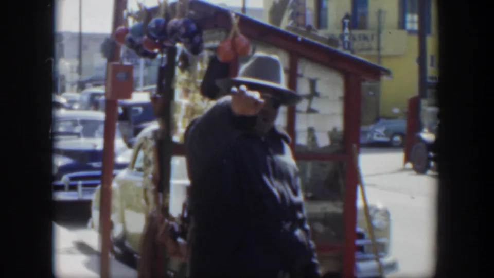 1951 Texas street frame showing a man in a dark jacket raising his hand near a red stall and parked cars.