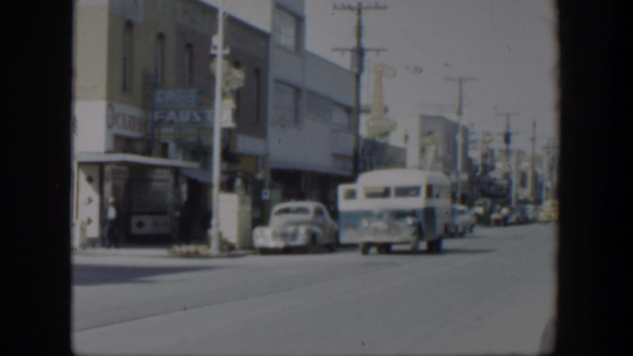 1951 Texas street view with a white bus, parked cars, storefront signage, and utility poles along a commercial block.