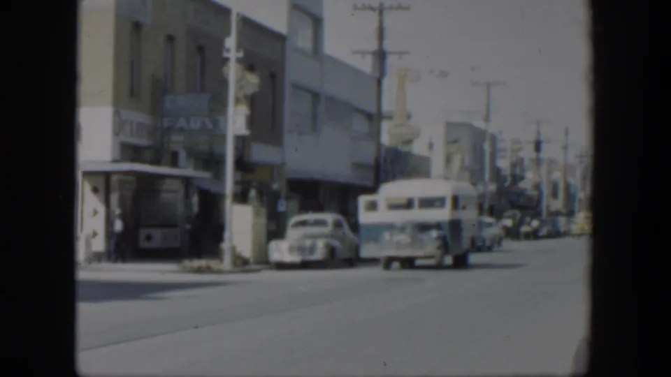 1951 Texas street view with a white bus, parked cars, storefront signage, and utility poles along a commercial block.