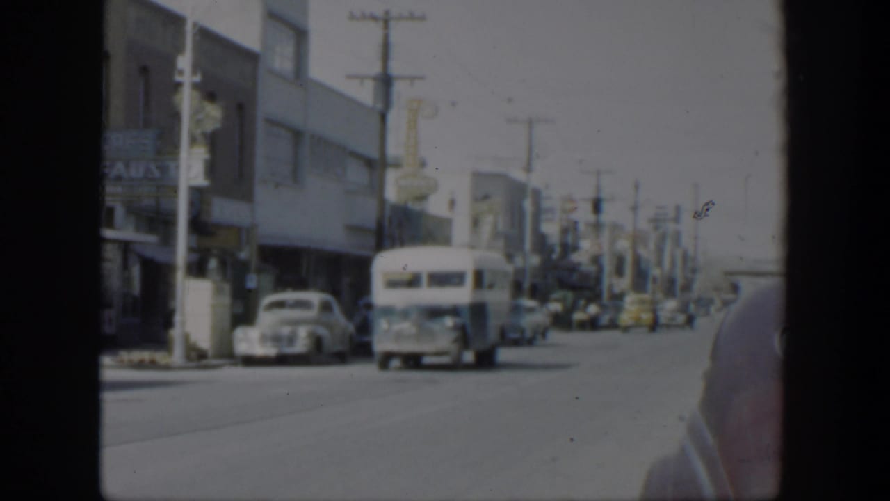 A white bus moves through a 1951 Texas commercial street scene with cars, signs, and roadside buildings visible.