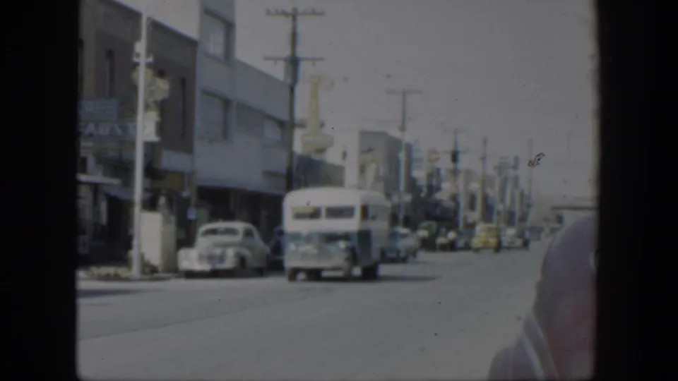 A white bus moves through a 1951 Texas commercial street scene with cars, signs, and roadside buildings visible.
