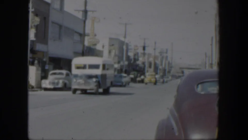 1951 Texas street frame showing a white bus nearer the camera, parked cars, storefront facades, and a car edge in the foreground.