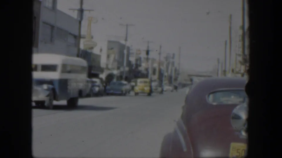 1951 Texas 8mm frame showing a sunlit commercial street with a white bus, parked cars, and a maroon sedan close to the moving camera.