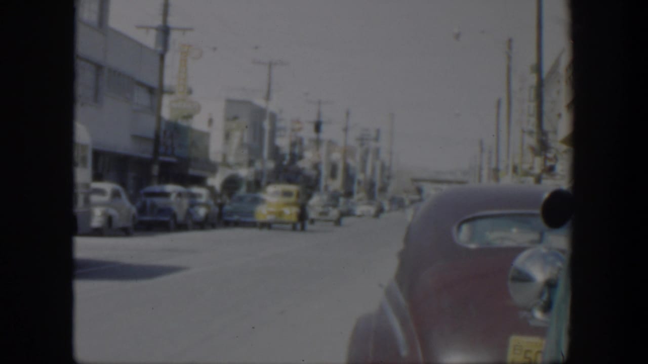1951 Texas street view from a moving vehicle, with parked mid-century cars, storefront buildings, overhead lines, and a maroon sedan near frame edge.