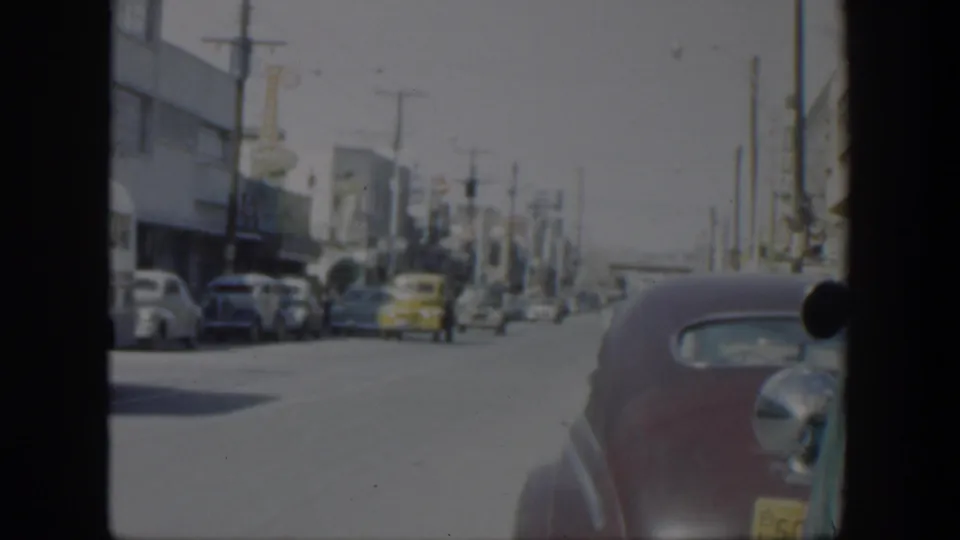 1951 Texas street view from a moving vehicle, with parked mid-century cars, storefront buildings, overhead lines, and a maroon sedan near frame edge.