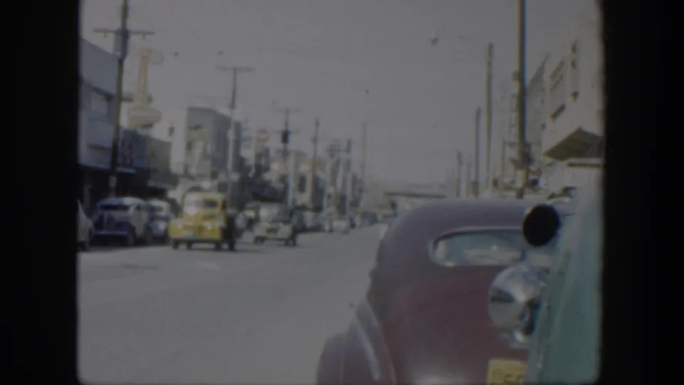 1951 Texas 8mm street frame with a yellow car driving ahead, rows of parked cars, storefront facades, and a maroon sedan close to camera.