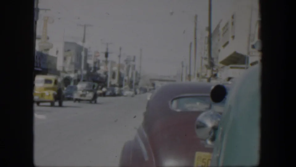 1951 Texas moving-car viewpoint showing a yellow car on the roadway, parked vehicles along storefronts, utility poles, and a maroon sedan in foreground.