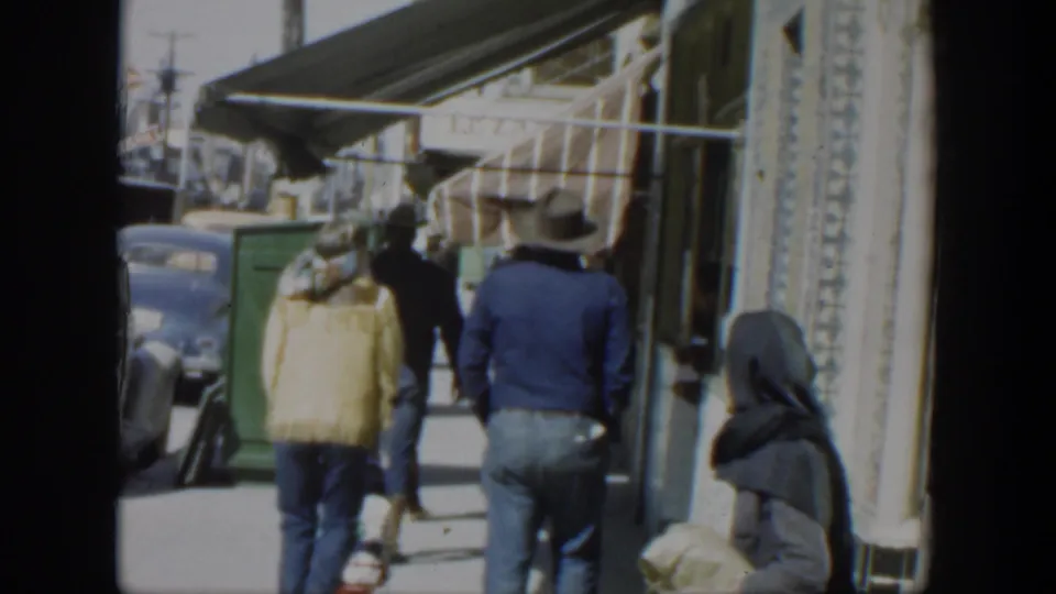 1951 Texas 8mm still with a headscarfed woman near the camera, pedestrians ahead, storefront awnings, and a car at the curb.